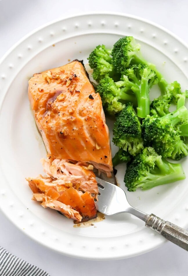 An overhead closeup photo of a filet of maple glazed salmon and steamed broccoli on a plate. A fork is flaking off some of the salmon.