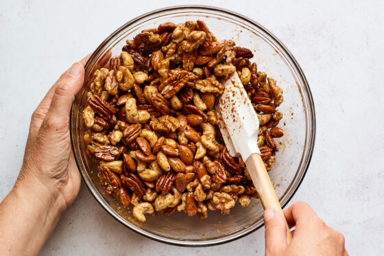 Walnuts, almonds, pecans and cashews being mixed with maple syrup, salt and cayenne pepper in a mixing bowl with a spatula.