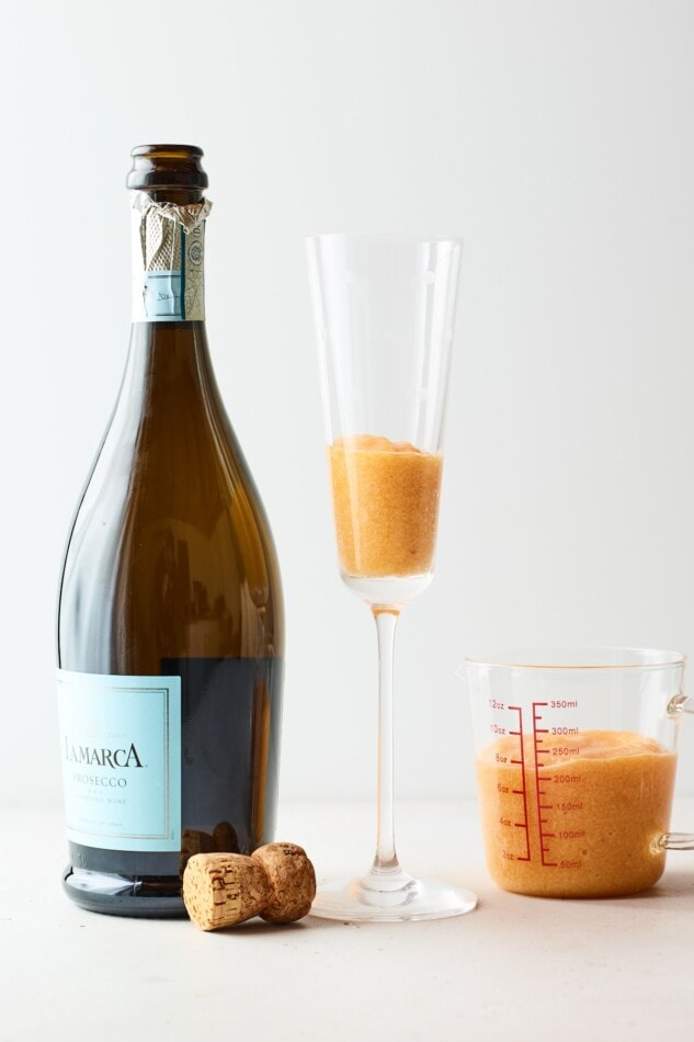 A champagne flute containing peach puree. A bottle of Prosecco is next to the glass but has not been poured into the glass yet.