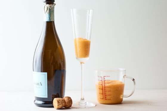 A champagne flute containing peach puree. A bottle of Prosecco is next to the glass but has not been poured into the glass yet.