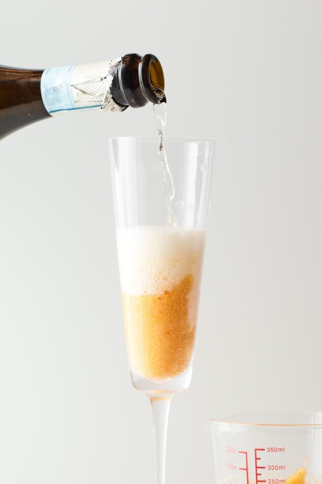 A closeup of a champagne flute containing peach puree. Prosecco is being poured into the glass from a bottle.