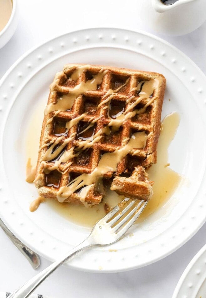 An overhead photo looking down at a plate with a rectangular protein waffle that has been drizzled with maple syrup and peanut butter. A fork rests on the plate with a piece of the waffle.