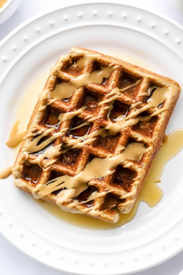 An overhead and close up photo looking at a plate with a rectangular protein waffle that has been drizzled with maple syrup and peanut butter.