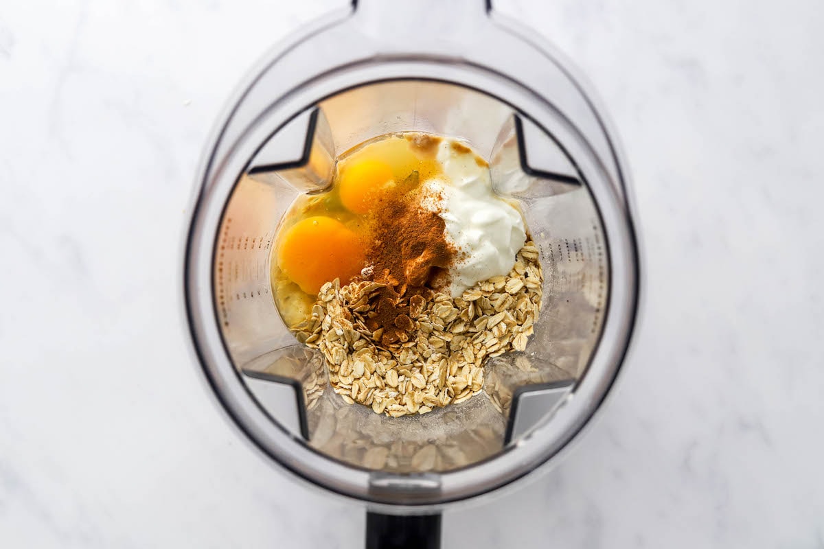 All ingredients for protein waffles in a high powered blender. Two eggs, greek yogurt, almond milk, maple syrup, vanilla extract, vanilla protein powder, rolled oats, baking powder, baking soda, cinnamon and sea salt.