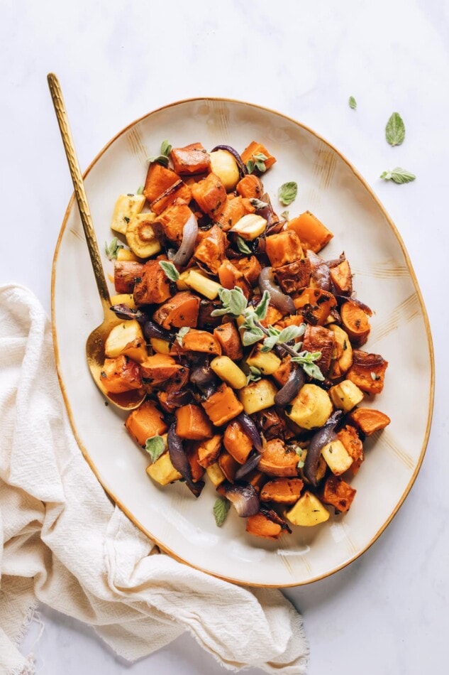 Caramelized roasted root vegetables with sweet potatoes, carrots, parsnips and fresh oregano. Such an easy side for the holidays or everyday meals. A serving platter with roasted root vegetables. A gold spoon rests in the platter.