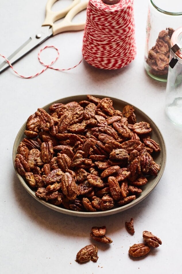 A shallow serving dish containing rosemary candied pecans. A spool of red and white striped twine and scissors are in the background.