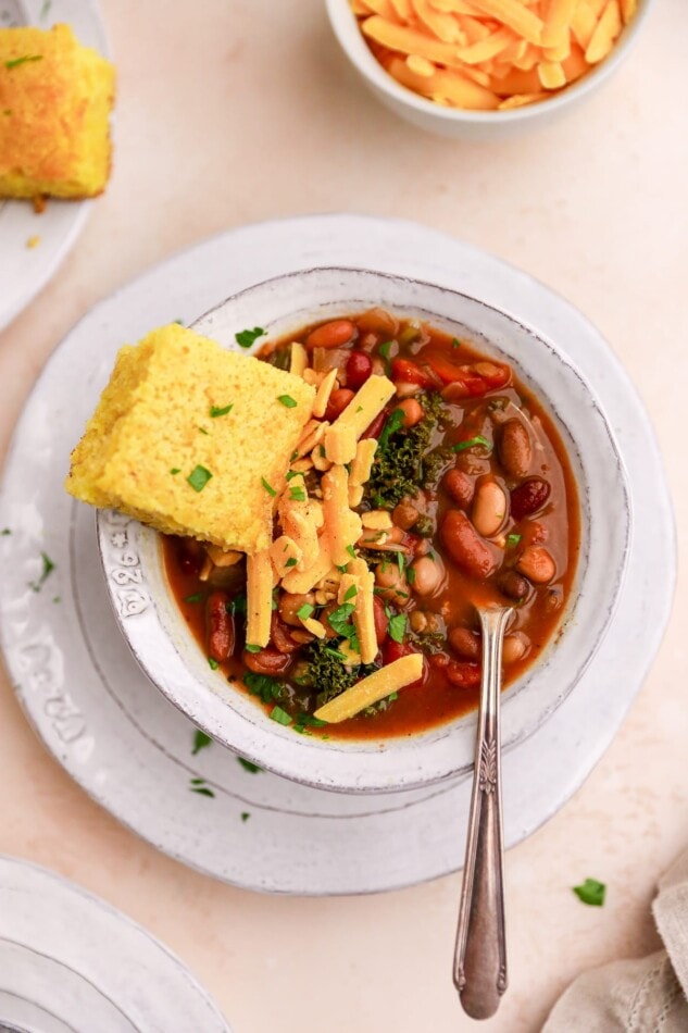 A bowl of 15 bean soup topped with a slice of corn bread, shredded cheese and fresh parsley. A spoon rests in the bowl.