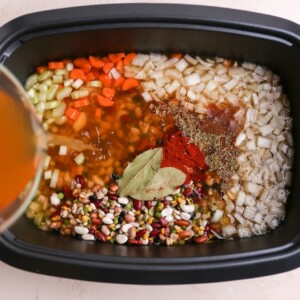Broth added to a slow cooker with bay leaves, spices, beans, oil, onion, garlic, carrots and celery.