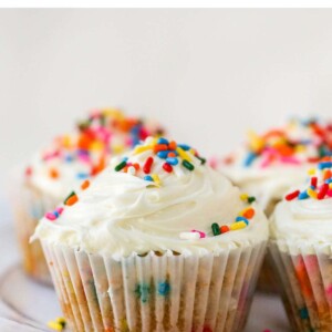 Four almond flour cupcakes wrapped in paper liners on a plate. The cupcakes have been topped with vanilla icing and rainbow sprinkles.