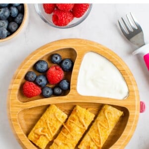 A plate with dividers. The largest portion has 3 slices of baby french toast. The 2 others hold yogurt and a mix of blueberries and raspberries. A small toddler fork is next to the plate.