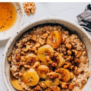 A bowl containing banana bread oatmeal that has been topped with caramelized bananas and walnuts. A spoon rests in the bowl.
