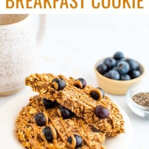 An oatmeal breakfast cookie on a plate that has been cut in half, the halves are resting on each other exposing the inside of the cookie. Behind the plate is a small container of chia seeds and blueberries as well as a mug of tea.