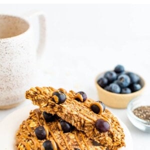 An oatmeal breakfast cookie on a plate that has been cut in half, the halves are resting on each other exposing the inside of the cookie. Behind the plate is a small container of chia seeds and blueberries as well as a mug of tea.