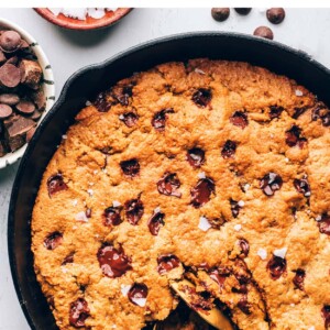 A cast iron skillet containing a chocolate chip cookie. Two spoons are removing a portion.