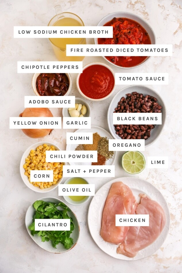Ingredients measured out to make chicken enchilada soup: chicken broth, fire roasted diced tomatoes, chipotle, adobo sauce, tomato sauce, yellow onion, garlic, black beans, cumin, oregano, chili powder, salt, pepper, lime, corn, olive oil, chicken and cilantro.