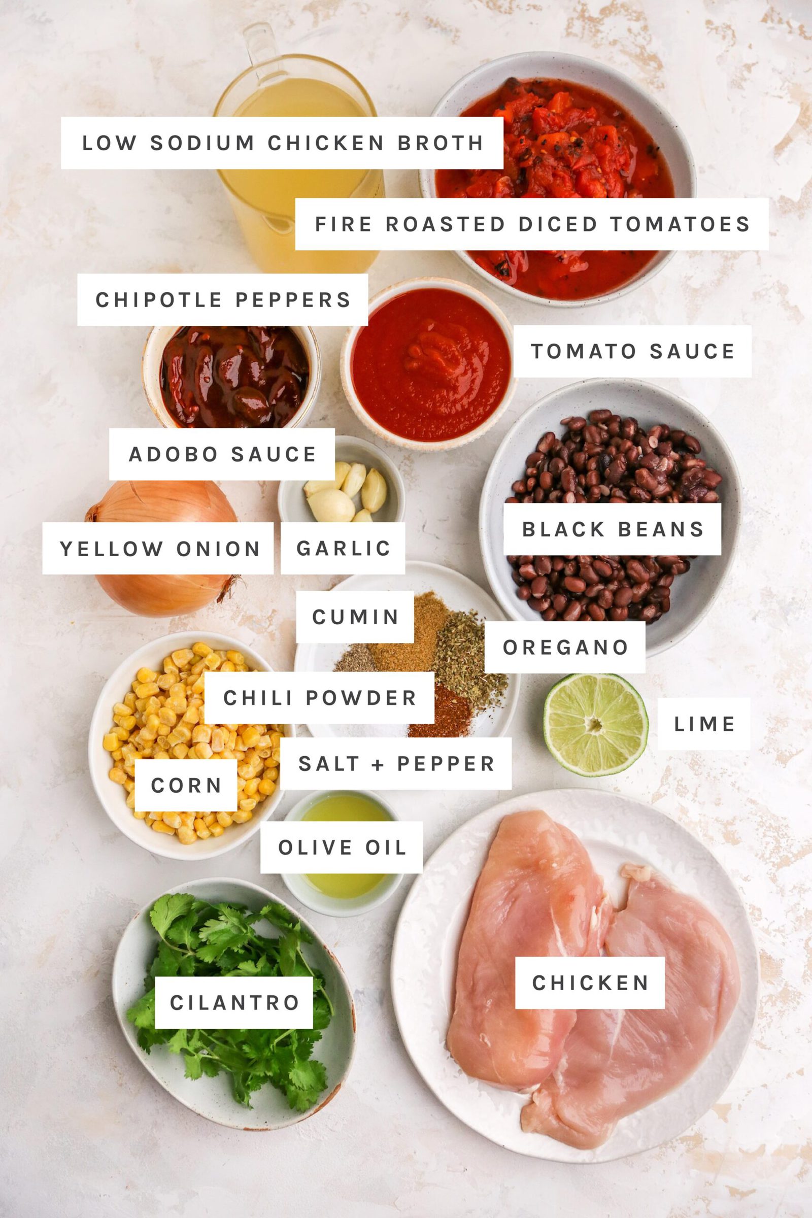 Ingredients measured out to make chicken enchilada soup: chicken broth, fire roasted diced tomatoes, chipotle, adobo sauce, tomato sauce, yellow onion, garlic, black beans, cumin, oregano, chili powder, salt, pepper, lime, corn, olive oil, chicken and cilantro.