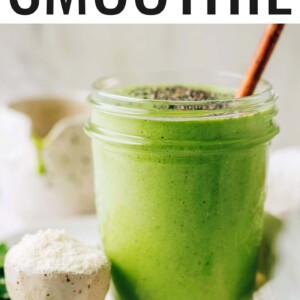 A mason jar containing green protein smoothie. The jar is resting on a plate with a small bowl of protein powder. The smoothie has been topped with chia seeds and a wooden straw sticks out of the top of the glass.