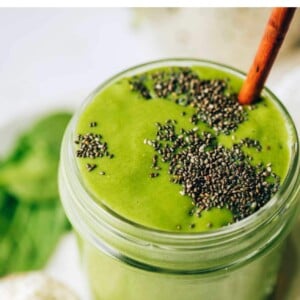 A mason jar containing green protein smoothie topped with chia seeds. A wooden straw rests inside the jar.