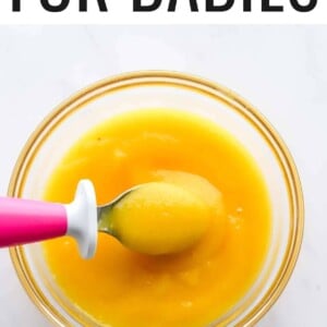 Bowl with mango puree baby food, and a baby spoon taking a bite.
