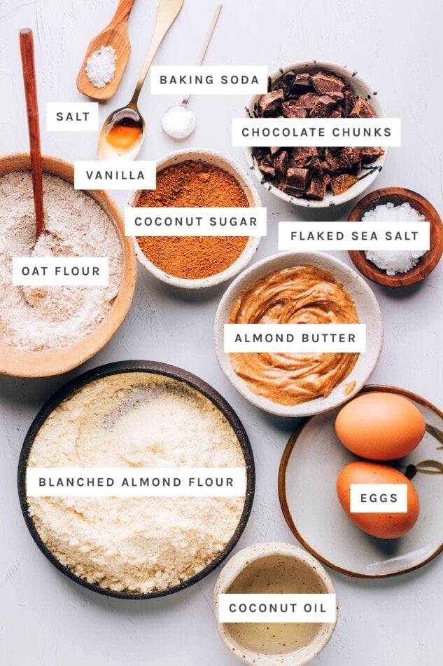 Ingredients measured out to make a chocolate chip cookie skillet: salt, vanilla, baking soda, chocolate chunks, coconut sugar, flaked sea salt, oat flour, almond butter, almond flour, eggs and coconut oil.