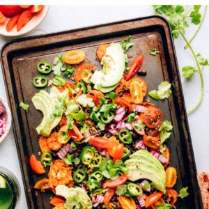 A hand scooping up a sweet potato round topped with black beans, cheese, avocado slices, and fresh cilantro.