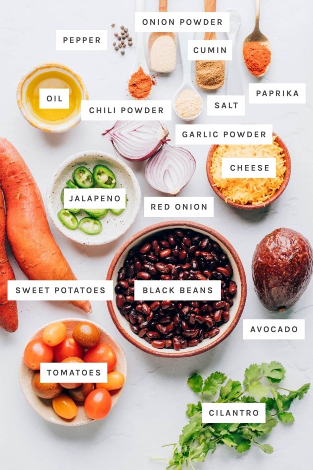 Ingredients measured out to make sweet potato nachos: pepper, onion powder, cumin, salt, paprika, oil, chili powder, garlic powder, cheese, red onion, jalapeno, sweet potatoes, black beans, avocado, tomatoes and cilantro.