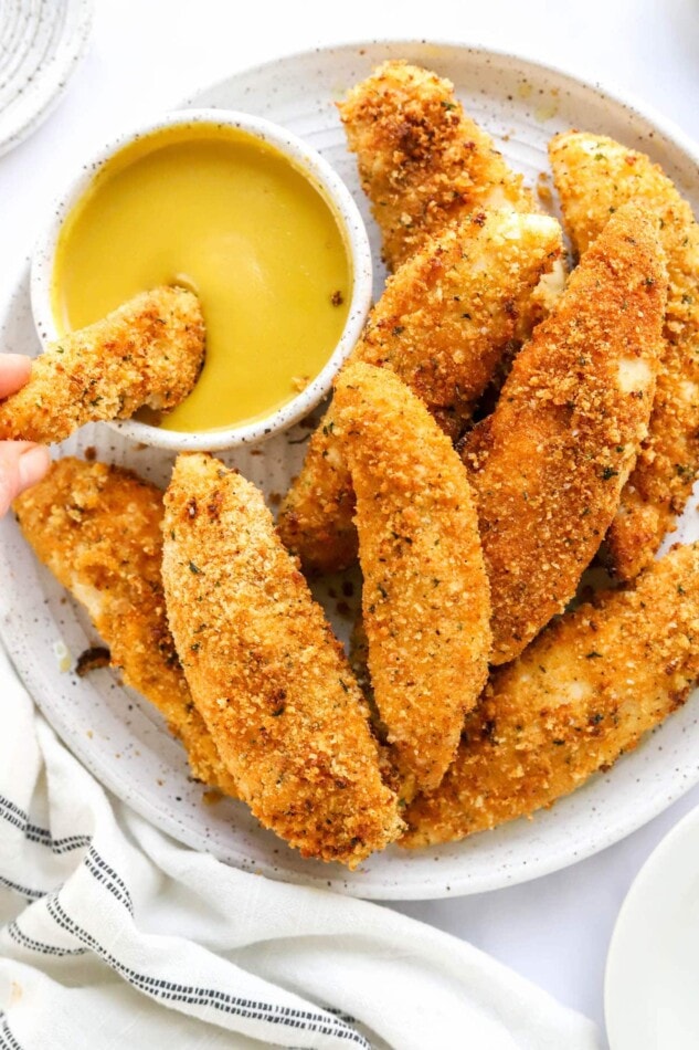 Chicken tenders on a plate with a bowl of dipping sauce. A hand is dipping one of the tenders into the sauce.