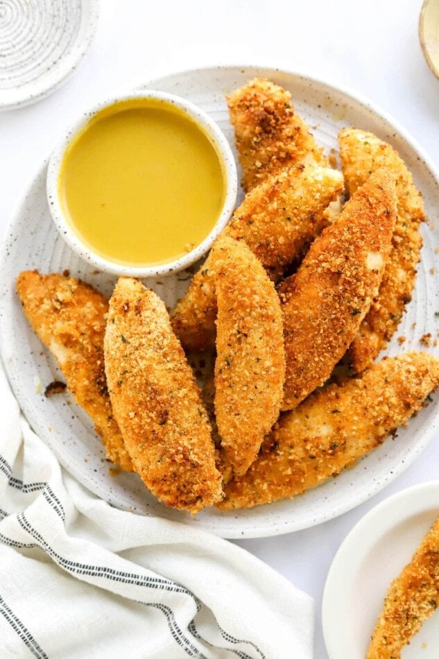 An overhead view of a plate holding air fryer chicken tenders. There is a bowl of honey mustard dipping sauce on the plate with the tenders.