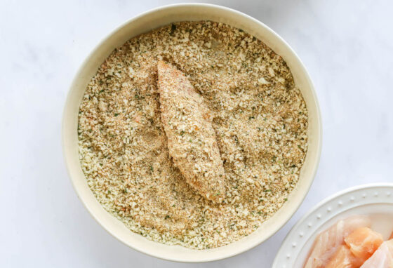 A chicken tender being dredged in seasoning after being coated in an egg wash.