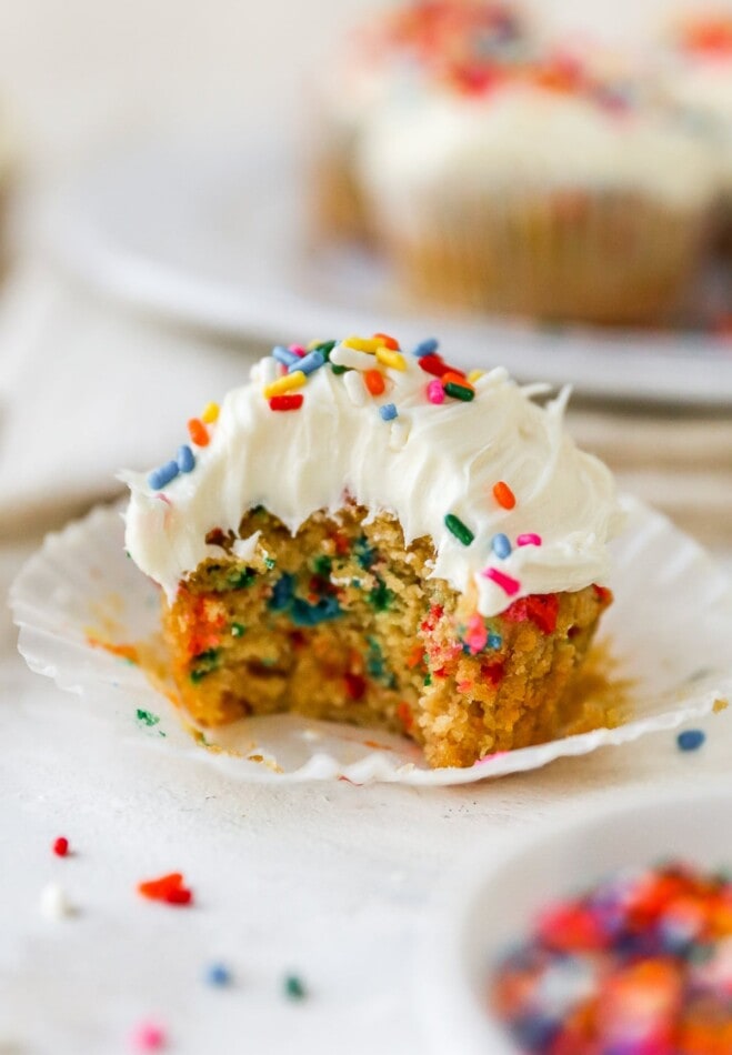 A cupcake topped with vanilla icing and rainbow sprinkles that has a bite removed from it.