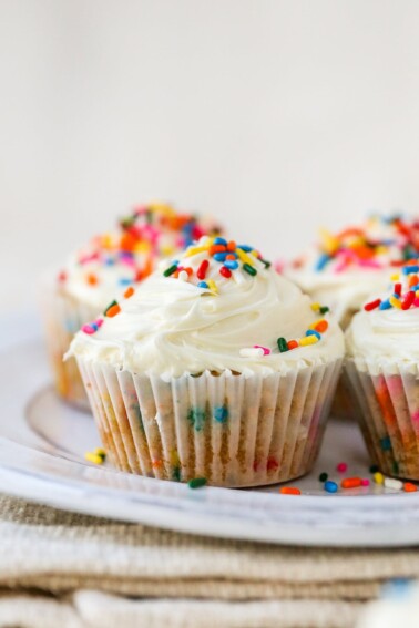 Four almond flour cupcakes wrapped in paper liners on a plate. The cupcakes have been topped with vanilla icing and rainbow sprinkles.