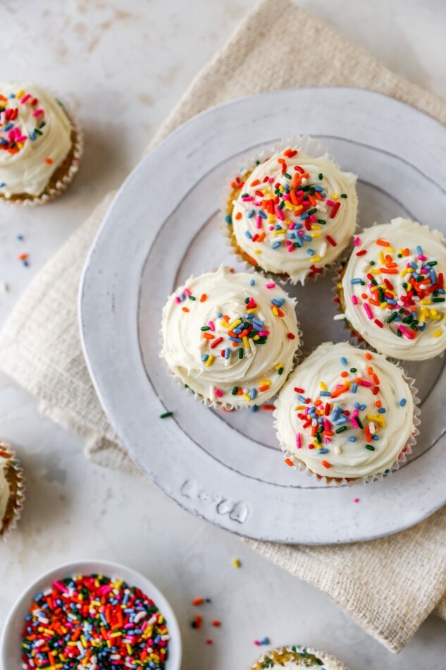 An overhead view of 4 cupcakes on a plate. There is a bowl of rainbow sprinkles nearby.