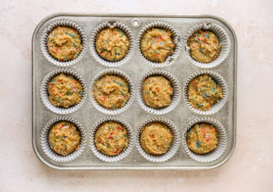 A 12-cavity cupcake tin with paper liners. Cupcake batter has been evenly distributed to all cavities.