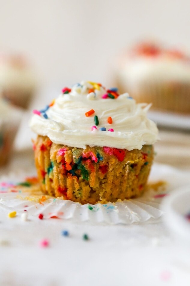 An almond flour cupcake that has been unwrapped from the paper liner. It is topped with vanilla icing and rainbow sprinkles.
