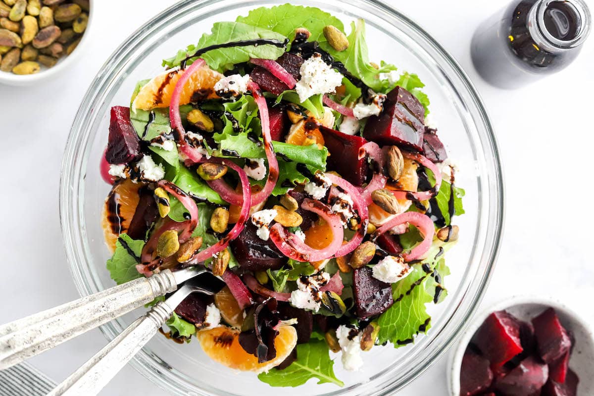 Ingredients for baby beet and clementine salad layered in a serving bowl. Serving utensils rest in the bowl.