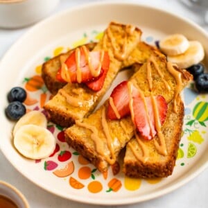 Slices of baby french toast on a plate topped with an almond butter drizzled and strawberry slices. There are banana and blueberry slices on the plate.