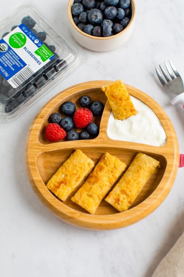 A divided plate for kids with 3 sections. The largest section has 3 slices of baby french toast. The two smaller sections holds an assortment of blueberries and raspberries and the other some yogurt. Another slice of baby french toast is being dipped into the yogurt.