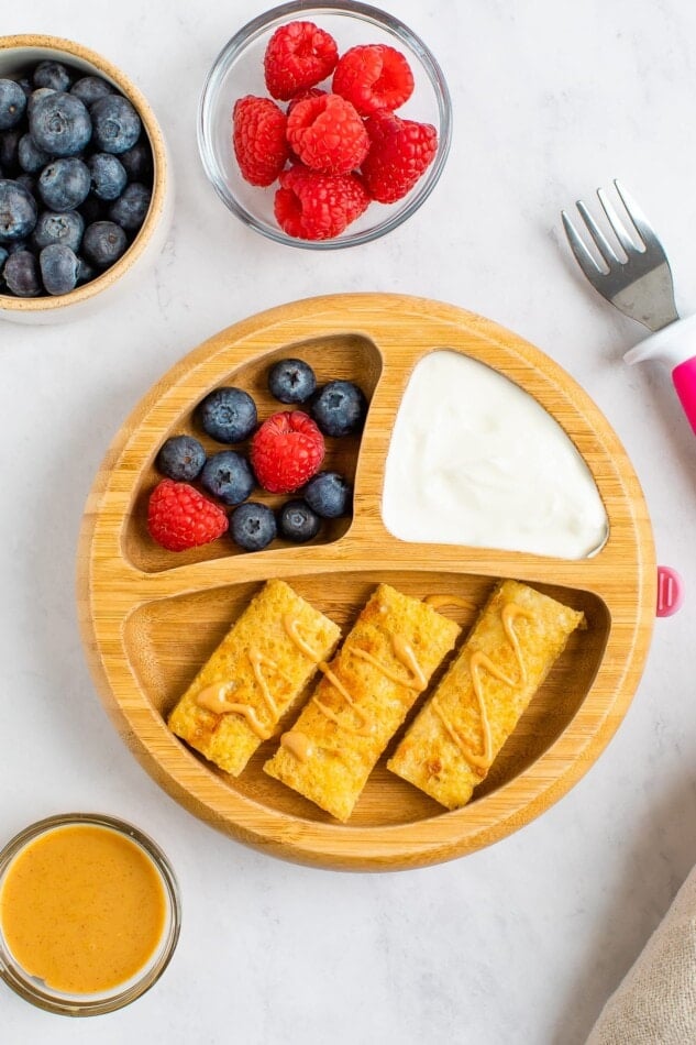 A plate with dividers. The largest portion has 3 slices of baby french toast. The 2 others hold yogurt and a mix of blueberries and raspberries. A small toddler fork is next to the plate.