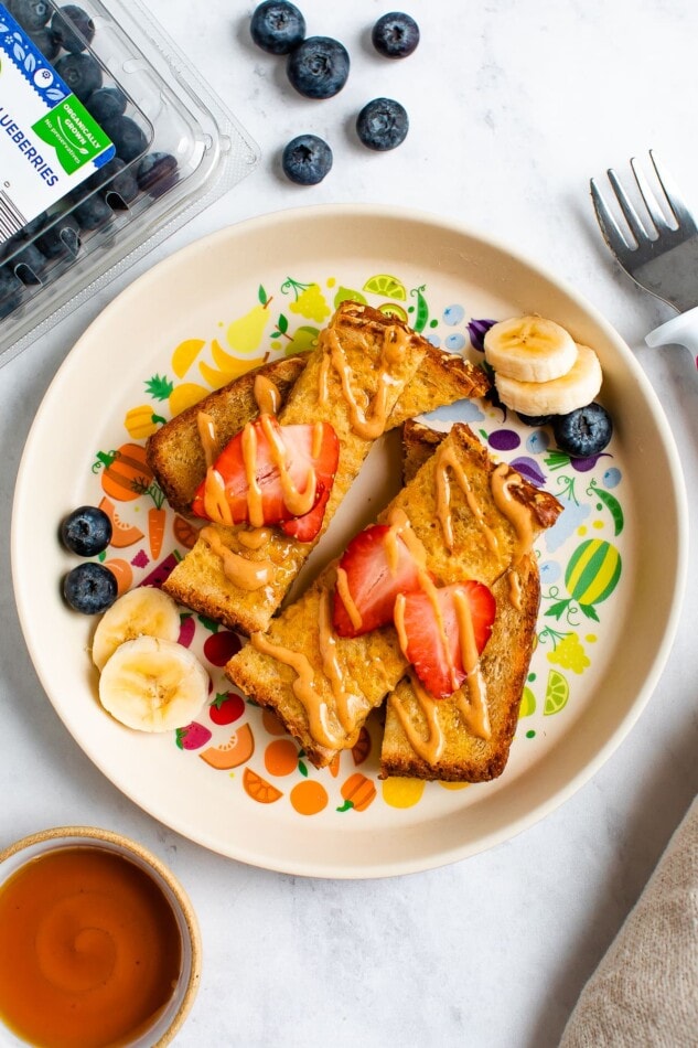 Slices of baby french toast on a plate topped with an almond butter drizzled and strawberry slices. There are banana and blueberry slices on the plate.