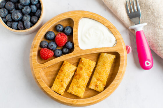 A plate with dividers. The largest portion has 3 slices of baby french toast. The 2 others hold yogurt and a mix of blueberries and raspberries. A small toddler fork is next to the plate.