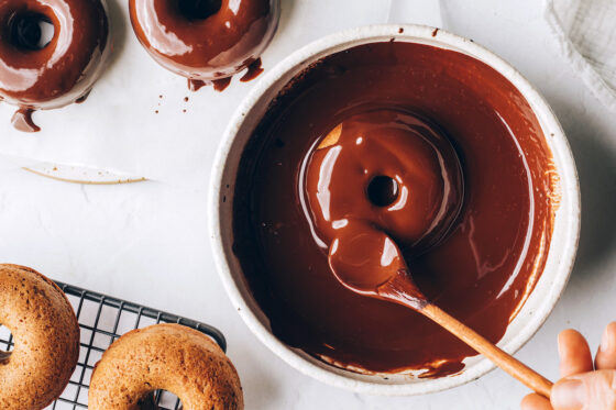 A donut in a bowl of chocolate sauce. A wooden spoon is covering the donut with chocolate.