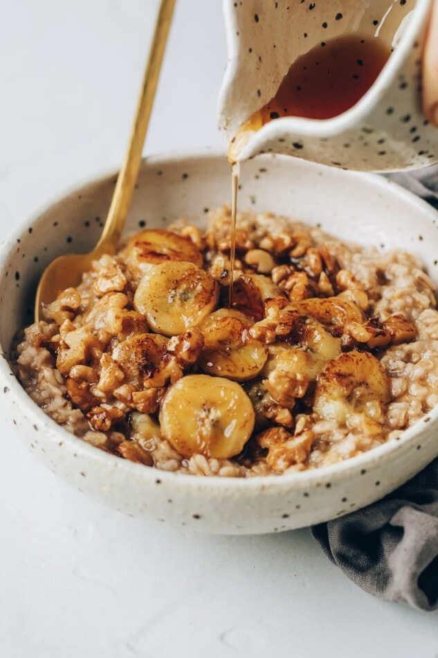 A bowl of banana bread oatmeal, topped with banana slices. A carafe is drizzling maple syrup over top of the oatmeal.