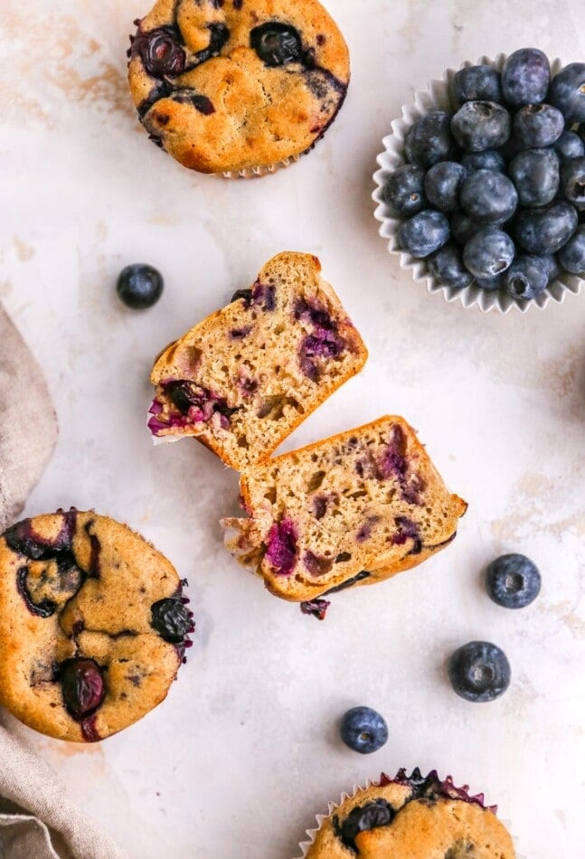 A greek yogurt muffin with blueberries sliced in half and laying on their sides, exposing the insides. Whole muffins are scattered around.