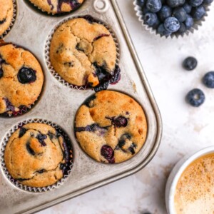 Six muffins in a muffin tin. A cup of blueberries is nearby.