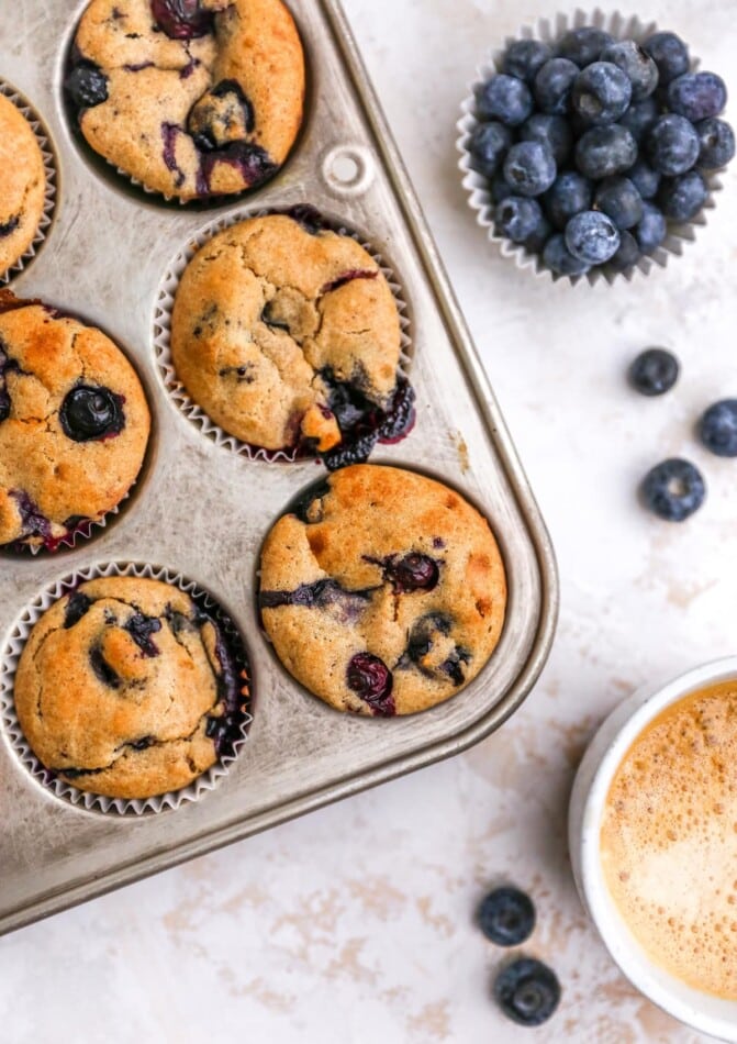 Six muffins in a muffin tin. A cup of blueberries is nearby.