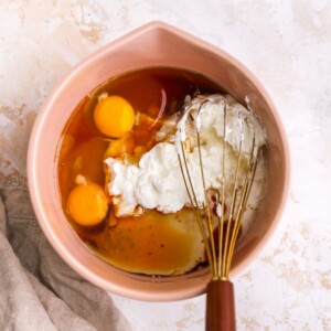 Eggs, Greek yogurt, oil, maple syrup, almond milk and vanilla extract in a medium bowl being mixed with a whisk.