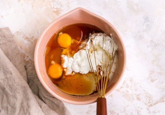 Eggs, Greek yogurt, oil, maple syrup, almond milk and vanilla extract in a medium bowl being mixed with a whisk.