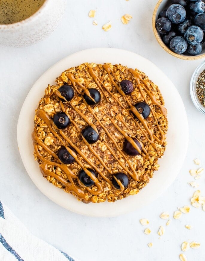 An overhead photo of a small plate with an oatmeal breakfast cookie on top. A small container of blueberries and chia seeds are next to the plate as well as a mug of tea.