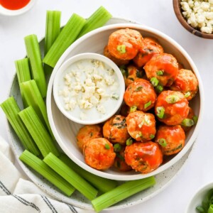 An overhead view of a bowl of buffalo chicken meatballs. A small bowl of blue cheese dip rests in the bowl with the meatballs and that bowl is on a plate surrounded by celery sticks.