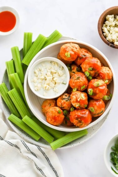 An overhead view of a bowl of buffalo chicken meatballs. A small bowl of blue cheese dip rests in the bowl with the meatballs and that bowl is on a plate surrounded by celery sticks.